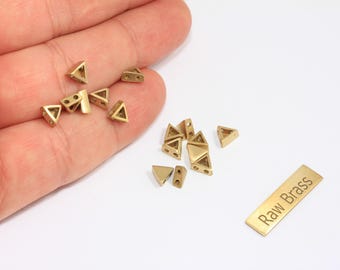 May include: A collection of small, golden-colored brass triangle beads, some with holes, scattered on a white surface. A rectangular brass tag with the text "Raw Brass" is also present. These beads are ideal for jewelry making.