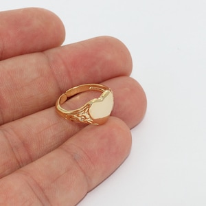 Peut inclure: Une bague dorée avec une face en forme de cœur. La bague a un anneau réglable et des détails décoratifs sur les côtés. La face en forme de cœur est de couleur crème clair. Bijoux.