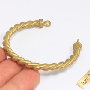 Op de afbeelding: Een goudkleurige, gedraaide messing armband met een lus sluiting. De armband heeft een touwachtig ontwerp en wordt op een witte achtergrond getoond. Een klein messing label met de tekst "Raw Brass" is ook zichtbaar.