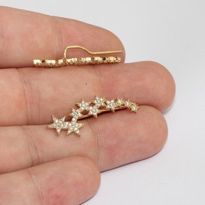 May include: Gold-coloured ear climbers with a star design. The earrings are embellished with small, clear crystals, creating a sparkling effect. The design includes a curved bar with multiple stars and a straight post for secure wear.