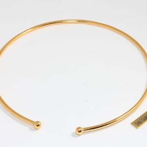 May include: A gold-plated choker necklace with a simple, elegant design. The necklace is a smooth, curved band with small ball ends. A small rectangular tag next to the necklace reads "24k Gold Plated".