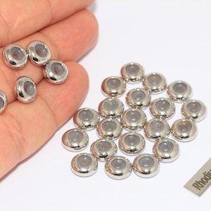 Pode incluir: Uma coleção de contas prateadas brilhantes com um centro cinzento. As contas são ovais e parecem ser de metal. Uma pequena etiqueta retangular com o texto "Rhodium Plated" também está presente.