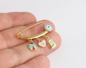 Puede incluir: Broche de alfiler de seguridad dorado con colgantes. Los colgantes incluyen una mariquita, un corazón rosa con una pequeña gema y un ojo azul. La mariquita tiene detalles en azul, rojo y dorado. El dije de ojo es azul claro.