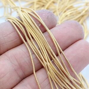 Può includere: Primo piano di un mucchio di cordoncino color oro, probabilmente per la creazione di gioielli o lavori artigianali. Il cordoncino è sottile, flessibile e ha una superficie strutturata. Il cordoncino è tenuto nella mano di una persona su sfondo bianco.