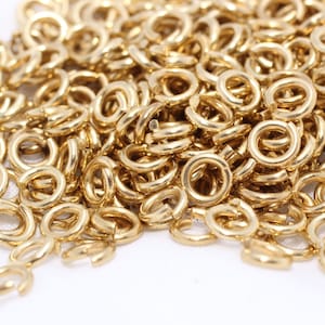 Könnte beinhalten: Nahaufnahme zeigt einen Haufen kleiner, goldfarbener Sprungringe. Die Ringe sind kreisförmig mit einer kleinen Öffnung und auf einer weißen Oberfläche verstreut. Diese Schmuckzubehörteile werden zum Verbinden von Schmuckkomponenten verwendet.