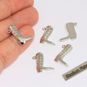 May include: Silver-colored cowboy boot charms with intricate detailing. The charms have a small loop at the top for attaching to jewelry. A small rectangular tag reads "Rhodium Plated".