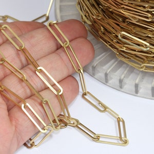 Puede incluir: Se muestra una cadena dorada de clip, un componente de joyería. La cadena está hecha de eslabones rectangulares, con una parte de la cadena extendida y el resto enrollado en una bobina. La cadena se muestra sobre un fondo blanco.