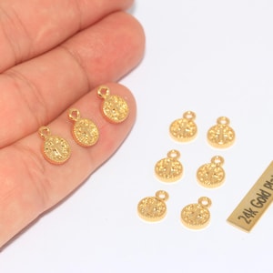 K&ouml;nnte beinhalten: Eine Sammlung kleiner, goldfarbener, runder Anh&auml;nger mit strukturierter Oberfl&auml;che und einer Schlaufe oben. Die Anh&auml;nger sind auf einer wei&szlig;en Oberfl&auml;che verstreut, einige werden in einer Hand gehalten. Ein kleines rechteckiges Etikett lautet "24k Gold Plated".
