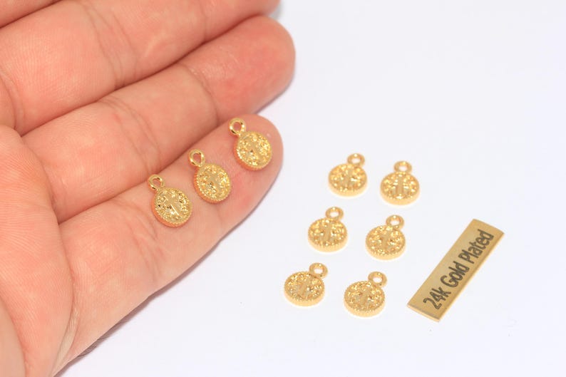 K&ouml;nnte beinhalten: Eine Sammlung kleiner, goldfarbener, runder Anh&auml;nger mit strukturierter Oberfl&auml;che und einer Schlaufe oben. Die Anh&auml;nger sind auf einer wei&szlig;en Oberfl&auml;che verstreut, einige werden in einer Hand gehalten. Ein kleines rechteckiges Etikett lautet "24k Gold Plated".