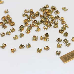 May include: A collection of small, gold-colored metal crimp covers scattered across the surface. Each piece features a rounded, hollow shape with a small loop at the top. A rectangular metal tag with the text RAW BRASS is visible in the corner.