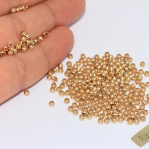 Può includere: Un insieme di piccole perline dorate, alcune tenute in mano, con una piccola etichetta rettangolare con la scritta "Raw Brass". Le perline sono sparse su una superficie bianca, suggerendo il loro utilizzo nella creazione di gioielli o nell'artigianato.