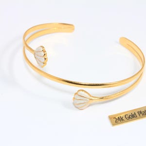 May include: Gold-plated cuff bracelet with white seashell-shaped accents. The open design features two parallel bands that curve around the wrist. A small rectangular tag reads "24k Gold Plated".