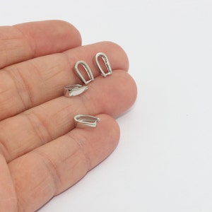 May include: Close-up of several small, silver-colored jewelry bails. The bails are in a U-shape with a small loop at the top for attaching to a chain or other jewelry component. The bails are on a light background.