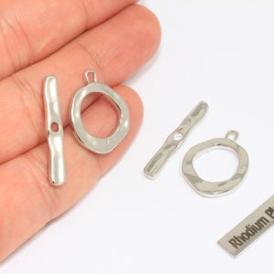 Puede incluir: Cierres de palanca plateados con textura martillada. El juego incluye dos barras de palanca, dos anillos circulares y una pequeña etiqueta rectangular que dice "Rhodium Plated". Estos hallazgos de joyería son ideales para collares y pulseras.