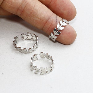 May include: Three silver leaf-design rings. Two rings are on a white surface, and one is on a finger. The rings have an open design, allowing for size adjustment. The leaf pattern wraps around the ring.