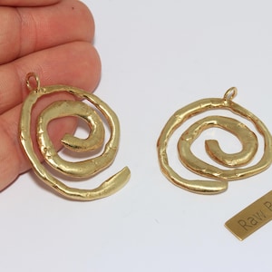 Può includere: Due ciondoli a spirale in ottone grezzo con finitura strutturata. Ogni ciondolo ha un anello nella parte superiore per essere appeso. È presente anche una piccola etichetta rettangolare in ottone con la scritta "Raw Brass".