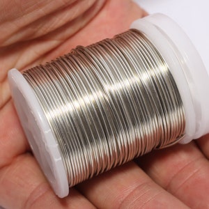 Puede incluir: Una mano sostiene una bobina de alambre brillante de color plateado. El alambre está enrollado alrededor de una bobina de plástico blanco. El alambre parece ser metálico y refleja la luz. La bobina se utiliza probablemente para la fabricación de joyas o manualidades.