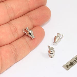 May include: Three silver-coloured miniature amphora charms, each with a small handle and base. One charm is held in a hand, with two others and a small metal tag that reads "Rhodium Plated" on a white surface.