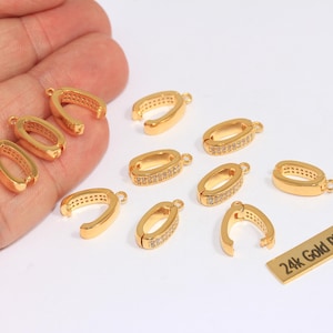 May include: Gold-plated jewelry clasps in various shapes, including oval and U-shaped designs. Some clasps are adorned with small, clear gemstones. A small rectangular tag reads "24k Gold Plated".