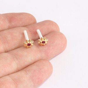 May include: A pair of gold-colored earrings with a flower design and a small, round, red gemstone in the center. The earrings have a white post. The earrings are displayed on a light background.