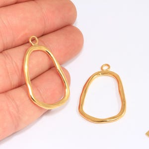 May include: Two gold-plated open-frame pendants with irregular, organic shapes. The pendants have a loop at the top for attaching to a chain or other jewelry component. A small rectangular tag reads "24k Gold Plated".