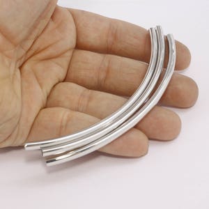 Könnte beinhalten: Mehrere gebogene, silberfarbene Metallröhren werden in einer Hand vor weißem Hintergrund gehalten. Die Röhren sind glatt und glänzend, mit einer abgerundeten Form. Das Bild ist gut beleuchtet und hebt den metallischen Glanz der Röhren hervor.
