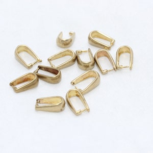 May include: A collection of gold-colored pendant bails, scattered on a white surface. The bails have a U-shaped design with a loop at the top for attaching to a chain or cord. These jewelry findings are ideal for creating necklaces and pendants.