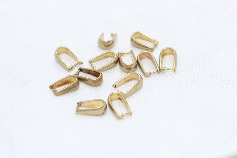 May include: A collection of gold-colored pendant bails, scattered on a white surface. The bails have a U-shaped design with a loop at the top for attaching to a chain or cord. These jewelry findings are ideal for creating necklaces and pendants.