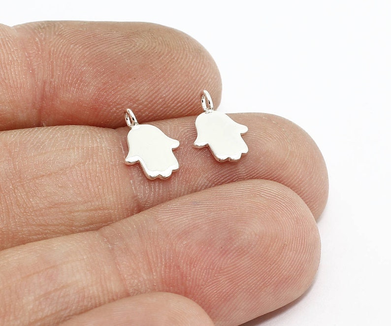 May include: Two small, silver-colored Hamsa hand charms. Each charm has a loop at the top for attaching to a chain or bracelet. The charms are smooth and shiny, with a simple, minimalist design. The background is white.