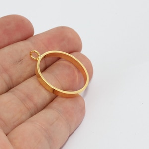Può includere: Un ciondolo ovale color oro con un piccolo anello nella parte superiore per una catena. Il ciondolo ha un profilo piatto e rettangolare e una superficie liscia e lucida. Il gioiello è mostrato su uno sfondo bianco.