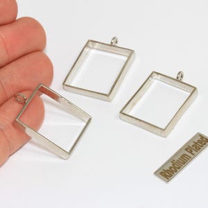 Puede incluir: Marcos de colgantes rectangulares plateados con un lazo para sujetar una cadena. La imagen también muestra una pequeña etiqueta metálica que dice "Rhodium Plated". Los colgantes están vacíos y listos para rellenar.
