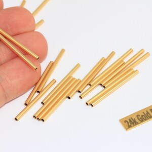 May include: A collection of small, gold-plated metal tubes, scattered on a white surface. The tubes are cylindrical with a hollow center. A small rectangular tag reads "24k Gold Plated".