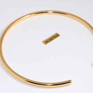 Könnte beinhalten: Eine goldbeschichtete Halskette mit offenem Design. Die Halskette hat eine helle, glänzende Goldfarbe und eine glatte, abgerundete Form. Ein kleines rechteckiges Etikett mit dem Text "24k Gold Plated" ist ebenfalls vorhanden.