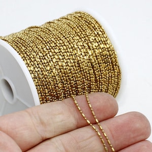 May include: A spool of gold-colored ball chain. The chain is made of small, elongated beads linked together. The spool is white and the chain is partially unraveled, showing the individual links. The background is white.
