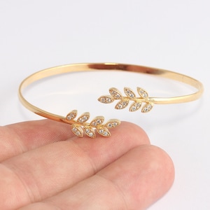 May include: A gold-colored open cuff bracelet with leaf-shaped accents. Each leaf features small, clear gemstones. The bracelet is set against a white background, highlighting the jewelry's design and details. The bracelet is a delicate piece of jewelry.