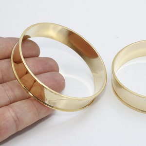 Pode incluir: Duas pulseiras abertas douradas. As joias são lisas e brilhantes, com uma superfície externa plana e bordas internas arredondadas. As pulseiras são exibidas em um fundo branco.