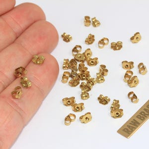 May include: A collection of small, gold-colored metal earring backs scattered on a white surface, with several resting on a palm. A rectangular metal tag with the text RAW BRASS is positioned near the earring backs, which are circular in shape.