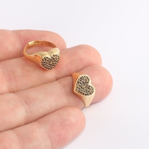 Puede incluir: Dos anillos dorados con caras en forma de corazón. Los corazones están llenos de pequeñas piedras oscuras. Un anillo está en un dedo, el otro está al lado. Fondo blanco.