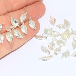 May include: Silver-colored leaf charms, perfect for jewelry making or crafting. The charms are detailed with veining and have a loop for easy attachment. Several charms are scattered on a white surface.
