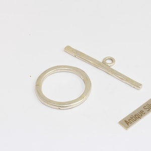 May include: An antique silver toggle clasp set, including a circular ring, a bar with a loop, and a rectangular tag with the text "Antique Silver". The clasp components are arranged on a white surface.