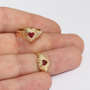 May include: Two gold rings with heart-shaped ruby-red center stones. The rings have a detailed design around the heart and band. The rings are displayed on a light background.