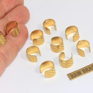 May include: A collection of ten small, open-ended gold-coloured metal cuffs, each featuring a diagonal striped texture and a single small hole at the top. A rectangular metal tag with the text RAW BRASS is included with the components.