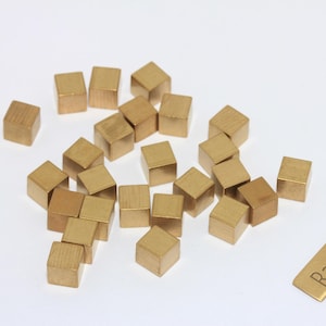 Può includere: Una collezione di piccoli cubi di ottone grezzo sparsi su una superficie bianca. È presente anche un'etichetta rettangolare in ottone con la scritta "Raw Brass". I cubi sono di un caldo colore dorato e l'estetica generale è minimalista.