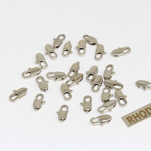 Pu&ograve; includere: Una collezione di piccoli moschettoni in metallo color argento sparsi su una superficie piana. Ogni chiusura &egrave; dotata di un meccanismo a molla per la creazione di gioielli. &Egrave; visibile un'etichetta metallica rettangolare con la parola RHODIUM.