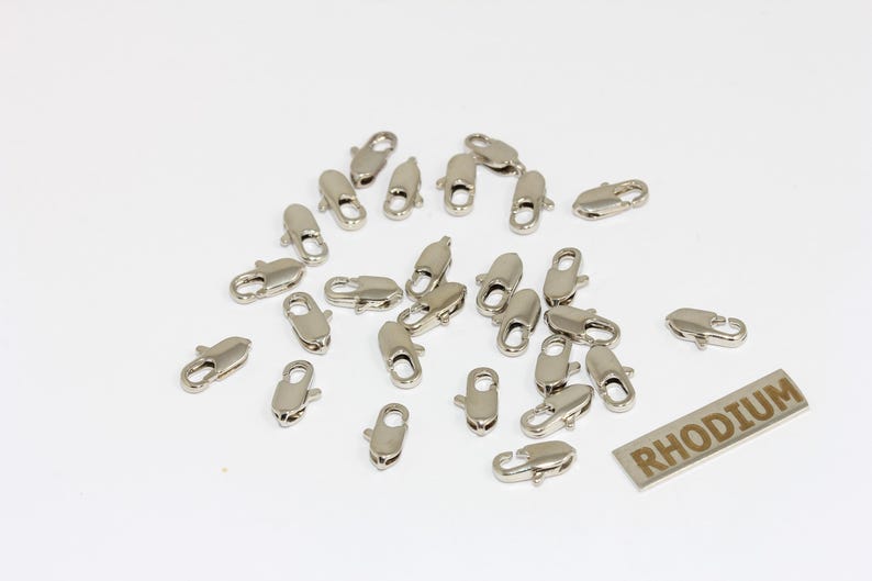 Pu&ograve; includere: Una collezione di piccoli moschettoni in metallo color argento sparsi su una superficie piana. Ogni chiusura &egrave; dotata di un meccanismo a molla per la creazione di gioielli. &Egrave; visibile un'etichetta metallica rettangolare con la parola RHODIUM.