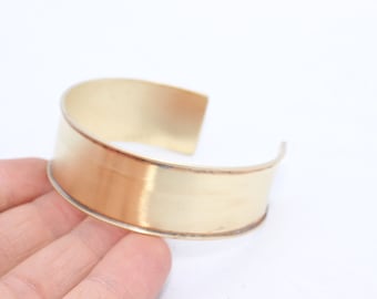 May include: A wide, open-ended cuff bracelet in a warm gold tone. The metal has a brushed finish and a slight curve, creating a simple yet elegant design. The bracelet is approximately 2 inches wide.