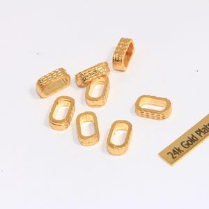 Puede incluir: Una colección de hallazgos de joyería rectangulares chapados en oro. Algunos tienen una textura tejida, mientras que otros son lisos. También hay una pequeña etiqueta dorada con el texto "24k Gold Plated". Los hallazgos están dispersos sobre una superficie blanca.
