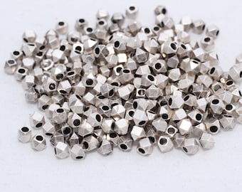 May include: A pile of small, silver-colored, faceted beads. Each bead has multiple sides and a hole running through the center. The beads are scattered on a white surface, creating a textured appearance.