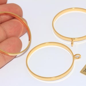 以下が含まれることがあります： ジュエリー作りに使用する、金メッキの丸いペンダントブランク。 画像には、3つの円形のフレームと「24k Gold Plated」と書かれた小さな長方形のタグが表示されています。 ペンダントは明るく光沢のある金色です。