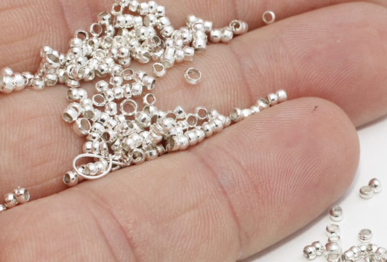 K&ouml;nnte beinhalten: Nahaufnahme von zahlreichen kleinen, silberfarbenen Perlen. Die Perlen sind zylindrisch mit abgerundeten Enden und einem zentralen Loch. Sie sind &uuml;ber eine Oberfl&auml;che verstreut, wobei einige zusammengefasst sind. Ideal f&uuml;r die Schmuckherstellung.